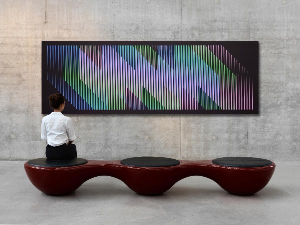 A woman seated on a modern, curved bench observes a large abstract artwork featuring colorful, diagonal lines and gradient hues against a black background, displayed on a concrete wall.